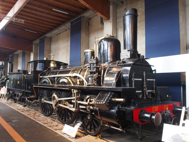Alte Dampflokomotive in einem Museums-Setting mit umgebenen Eisenbahnschienen, Informationsschildern und anderen Ausstellungsstücken unter einer beleuchteten Decke.