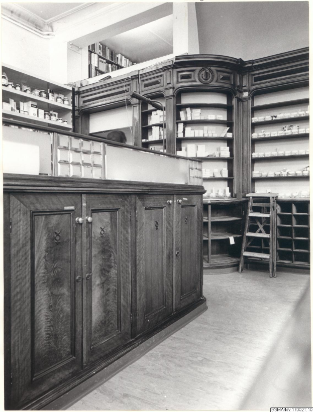 Ein Schwarz-Wei├č-Foto eines Apothekeninneren mit einer Theke, Schrankf├Ąchern, einer Leiter und Regalen mit verschiedenen Gegenst├Ąnden, mit Text ├╝ber dem Bild.