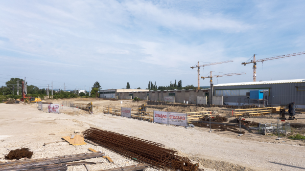 Baustelle mit verschiedenen Geräten wie Eisenstangen, hölzernen Gegenständen, bedruckten Platten, Pfählen, Drähten, Bäumen, Gebäuden und Kränen unter einem bewölkten Himmel.
