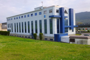 Ein großes weißes und blaues Gebäude thront auf einem saftigen grünen Feld, umgeben von Bäumen und Pflanzen, mit Fahrzeugen, Masten, Lampen, Schildern und Bergen im Hintergrund unter einem klaren blauen Himmel.