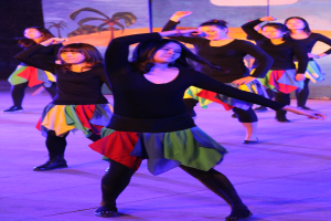 Junge Frauen in bunten Röcken und Schuhen tanzen energisch auf einer Bühne mit einem Wandgemälde im Hintergrund.