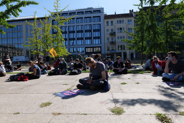 Eine Gruppe von Menschen sitzt vor einem Gebäude auf dem Boden während einer Demonstration in Berlin, einige tragen Masken und es liegen Gegenstände verstreut herum, unter einem klaren blauen Himmel mit Bäumen und Gebäuden im Hintergrund.