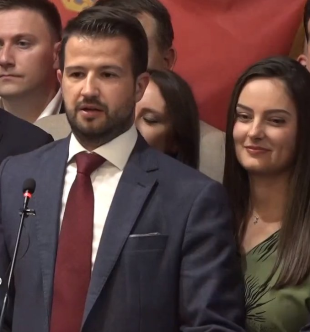 Ein Mann im Anzug spricht in ein Mikrofon auf einem Podium, mit einem Banner im Hintergrund und einer Menge davor, wahrscheinlich zu einem Regierungsverbot von sozialen Medien.