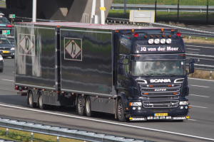 Ein schwarzer Sattelschlepper fährt auf einer Autobahn neben einer Brücke, mit anderen Fahrzeugen, Pfählen, Brettern und Gras im Hintergrund.
