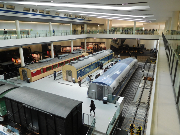 Eine großzügige Museumsausstellung zeigt verschiedene Züge in unterschiedlichen Größen und Farben, bei der Besucher vom Boden, Geländern und Säulen unter Deckenbeleuchtung zuschauen.