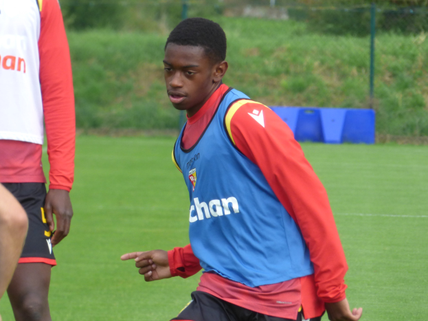 Ein junger Mann in einem blauen Hemd und schwarzen Shorts spielt Fußball auf einem grünen Feld, das von einem Zaun, Pflanzen und Bäumen umgeben ist.