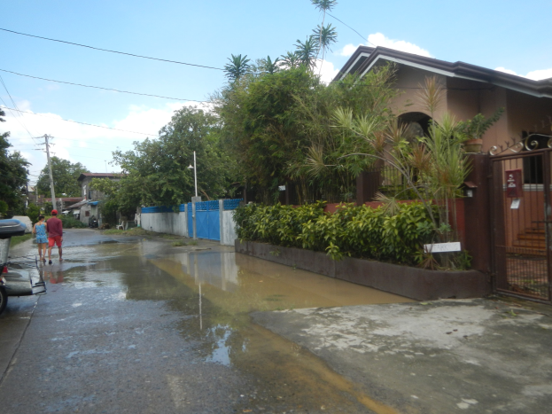 Eine überflutete Straße mit einem geparkten Motorrad, Menschen, die gehen, Gebäude, Bäume, Pflanzen, Pfosten, Drähte, ein Tor im Hintergrund und ein bewölkter Himmel mit einem Wasserzeichen.