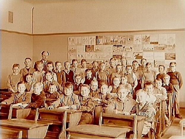 Ein Schwarz-Weiß-Foto einer Klasse mit Kindern auf Bänken und auf dem Boden sitzend und mit Papieren an der Rückwand.