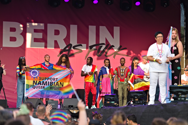 Gruppe von Menschen auf einer Bühne mit einem Banner 'Berlin as Fuck' mit Lautsprechern, Mikrofonen und einer feiernden Menge unten.
