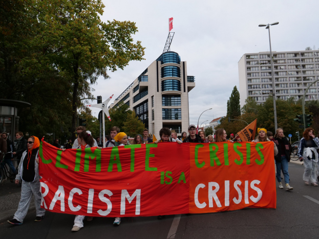 Gruppe von Menschen marschiert auf einer von Bäumen gesäumten Straße und hält ein Banner mit der Aufschrift "Klima-Krise ist eine Krise" hoch, mit Gebäuden und Laternenmasten im Hintergrund.