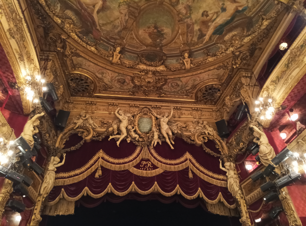 Theaterinnenraum mit roten Vorhängen, Kronleuchtern, Deckenmalerei und Skulpturen, mit einer Bühne und einigen Menschen unten.