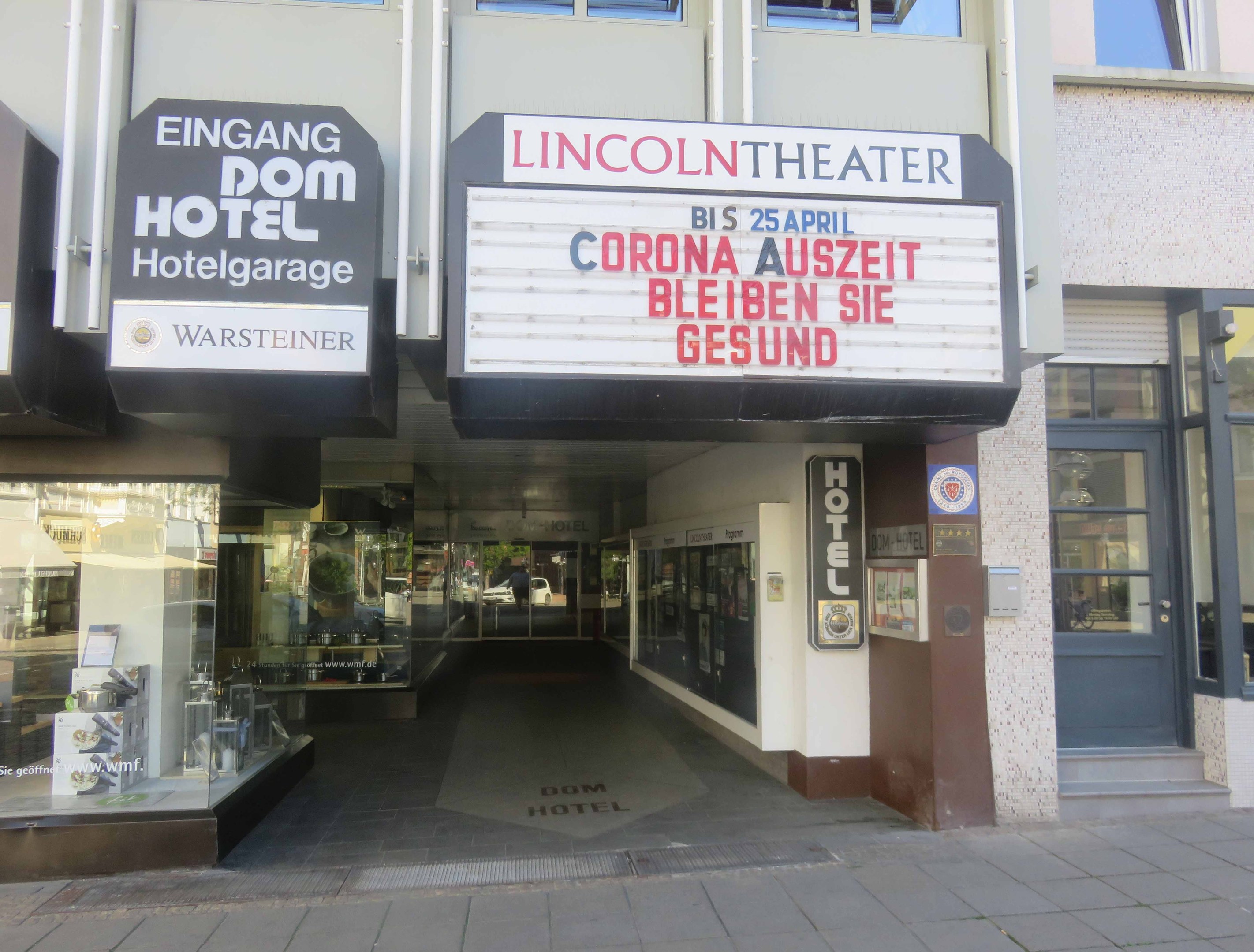 Außenansicht des Lincoln Theaters in Berlin, Deutschland, mit Glasfenstern und -türen sowie einer Tafel mit Text und einer belebten Stadtlandschaft im Inneren.