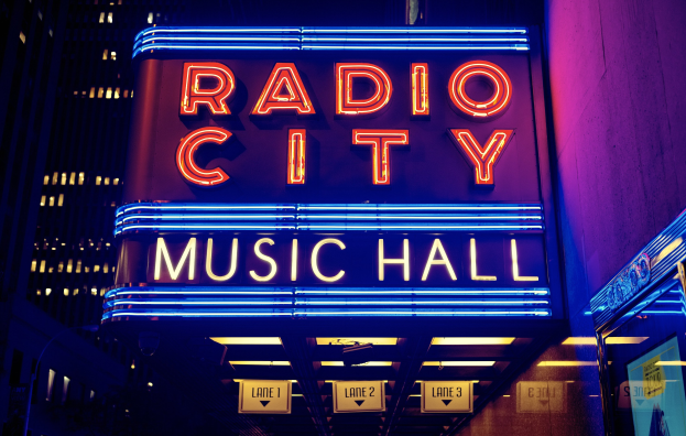 Radio City Music Hall in New York City, hell erleuchtet mit hohen Gebäuden im Hintergrund, mit einem "Radio City"-Schild und mehreren kleineren Schildern auf der rechten Seite.