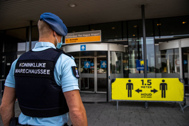 Ein Polizeibeamter in blauer Uniform und Mütze steht vor einem gläsernen Gebäude mit einem gelben Schild auf der rechten Seite und zusätzlichen Schildern im Hintergrund.