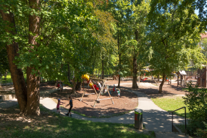 Ein Park mit einem Spielplatz in der Mitte, umgeben von Bäumen, mit Kindern auf der Ausrüstung, einem Mülleimer, Geländern, Pflanzen, Gras und Gebäuden im Hintergrund unter einem klaren blauen Himmel.