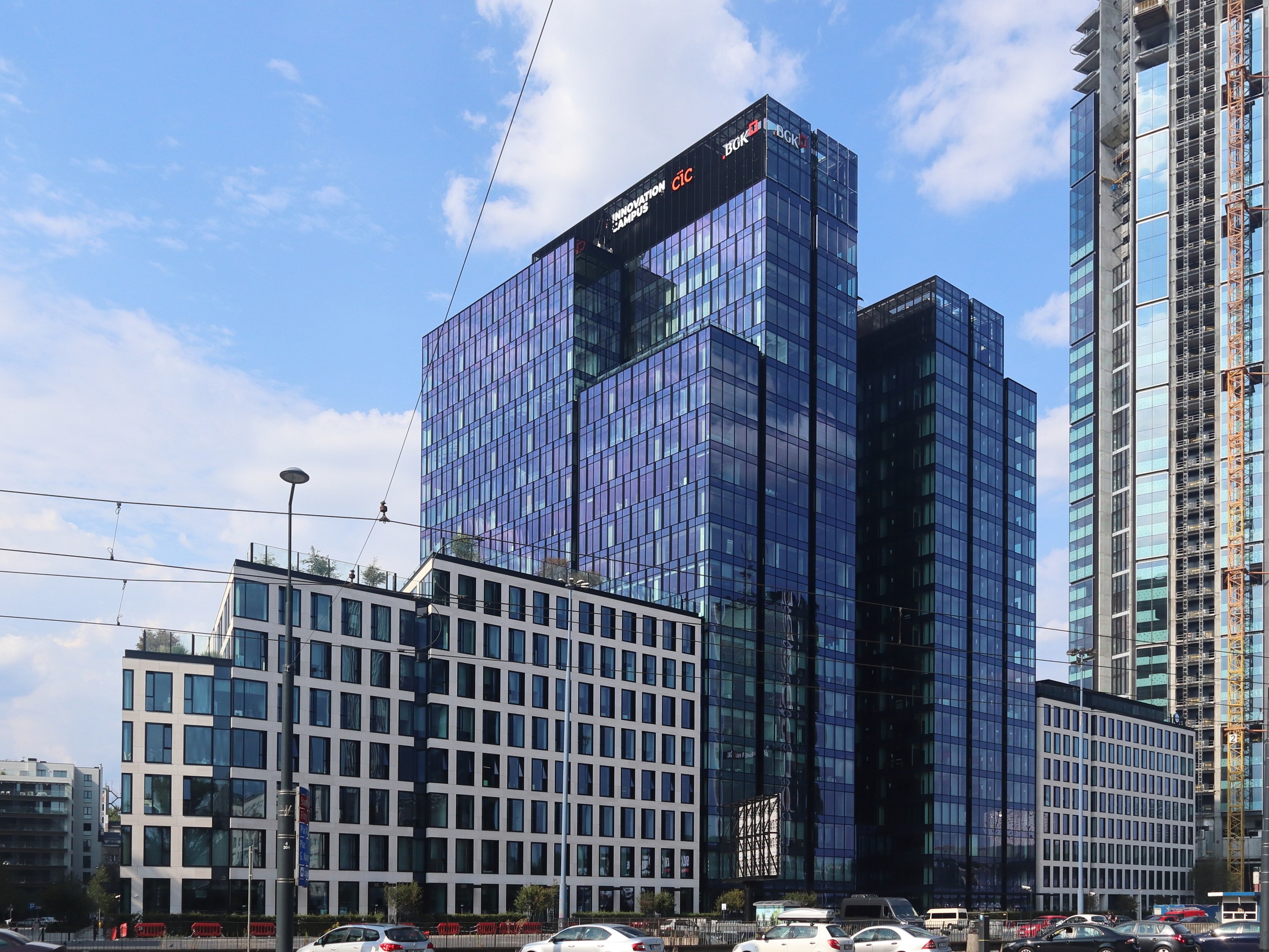 Hauptsitz der Deutschen Bank in Frankfurt, Deutschland, ein gro├čer mehrst├Âckiger Bau mit zahlreichen Fenstern, umgeben von urbaner Infrastruktur wie Stra├čenlaternen, elektrischen Kabeln, Fahrzeugen, Vegetation und einem bew├Âlktem Himmel.