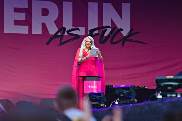 Frau in rosa Kleid, die in ein Mikrofon spricht, vor einem Publikum mit einem "Berlin as Fuck"-Plakat dahinter