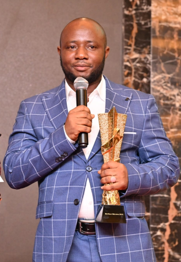 Ein Mann in einem blauen Anzug hält ein Mikrofon und eine Trophäe und steht selbstbewusst mit einem freudigen Gesichtsausdruck vor einer Wand im Hintergrund.