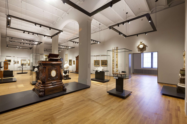 Ein großer Ausstellungsraum mit verschiedenen Kunstwerken, darunter Skulpturen, Glaskästen, gerahmten Stücken, einer Wanduhr, einem Fenster und Deckenleuchten, Teil der Nationalgalerie in Washington, D.C.