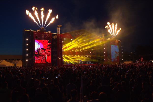 Eine große Menschenmenge auf einem Musikfestival schaut auf einer hell erleuchteten Bühne Performern zu, mit Zelten links und Feuerwerk in dunklem Himmel dahinter.
