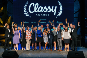 Eine Gruppe von Menschen auf einer Bühne, die strahlen und Auszeichnungen halten, mit Lautsprechern unten und einem Bildschirm im Hintergrund, der 'Classy Awards 2019' anzeigt.