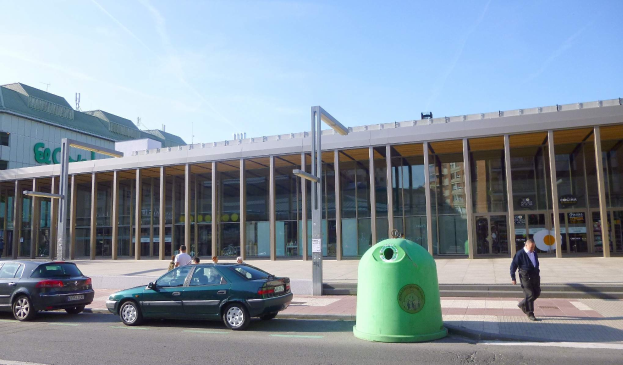 Grüner Müllcontainer auf einer Stadtstraße neben einem glaswandigen Gebäude mit Säulen, mit Fahrzeugen und Fußgängern im Hintergrund und einem bewölkten Himmel.