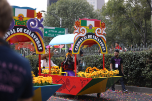 Eine Gruppe von Menschen auf einem blumen geschmückten Festwagen bei einem Umzug, umgeben von einem Metallzaun, Schildern, einer Straßenlaterne, einer Metallabsperrung, Pflanzen, Bäumen und einem Gebäude mit Fenstern unter einem bewölkten Himmel.