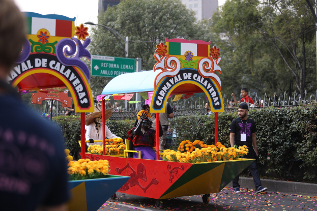 Eine Gruppe von Menschen auf einem blumen geschmückten Festwagen bei einem Umzug, umgeben von einem Metallzaun, Schildern, einer Straßenlaterne, einer Metallabsperrung, Pflanzen, Bäumen und einem Gebäude mit Fenstern unter einem bewölkten Himmel.