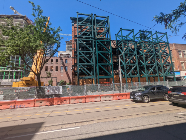 Große Metallbrücke im Bau an einer Baustelle mit parkenden Autos, einem Zaun, Bäumen, Gebäuden, einem Kran und einem Hintergrundhimmel.