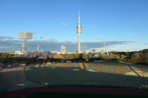 Außenansicht des Olympiastadions in Berlin, Deutschland, mit dem Fernsehturm (Fernsehturm) im Hintergrund, umgeben von B"umen, Geb"uden und beleuchteten Bereichen unter einem bew"olkten Himmel.