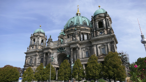 Der Berliner Dom, eine große Kathedrale mit Fenstern, Säulen, Bögen, Statuen und einem Turm, steht prominent in Berlin, Deutschland, mit Menschen in der Nähe, Bäumen, blühenden Pflanzen und einem bewölkten Himmel im Hintergrund.