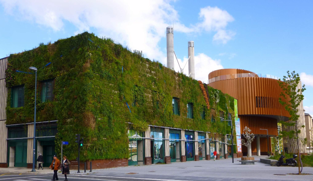 Ein großes Gebäude mit einer grünen Wand an der Seite, umgeben von Straßeninfrastruktur, Fahrzeugen, Fußgängern, Bäumen und einem bewölkten Himmel, als Teil des Green Building Project.