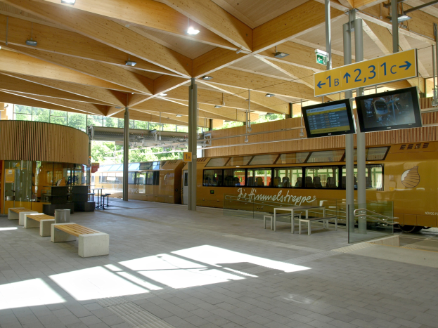 Ein belebter Bahnhof mit einem Zug auf den Schienen, umgeben von Bänken, Tischen, Pfählen, Schildern, Laternen und Bäumen, mit Menschen, die auf den Zug warten.