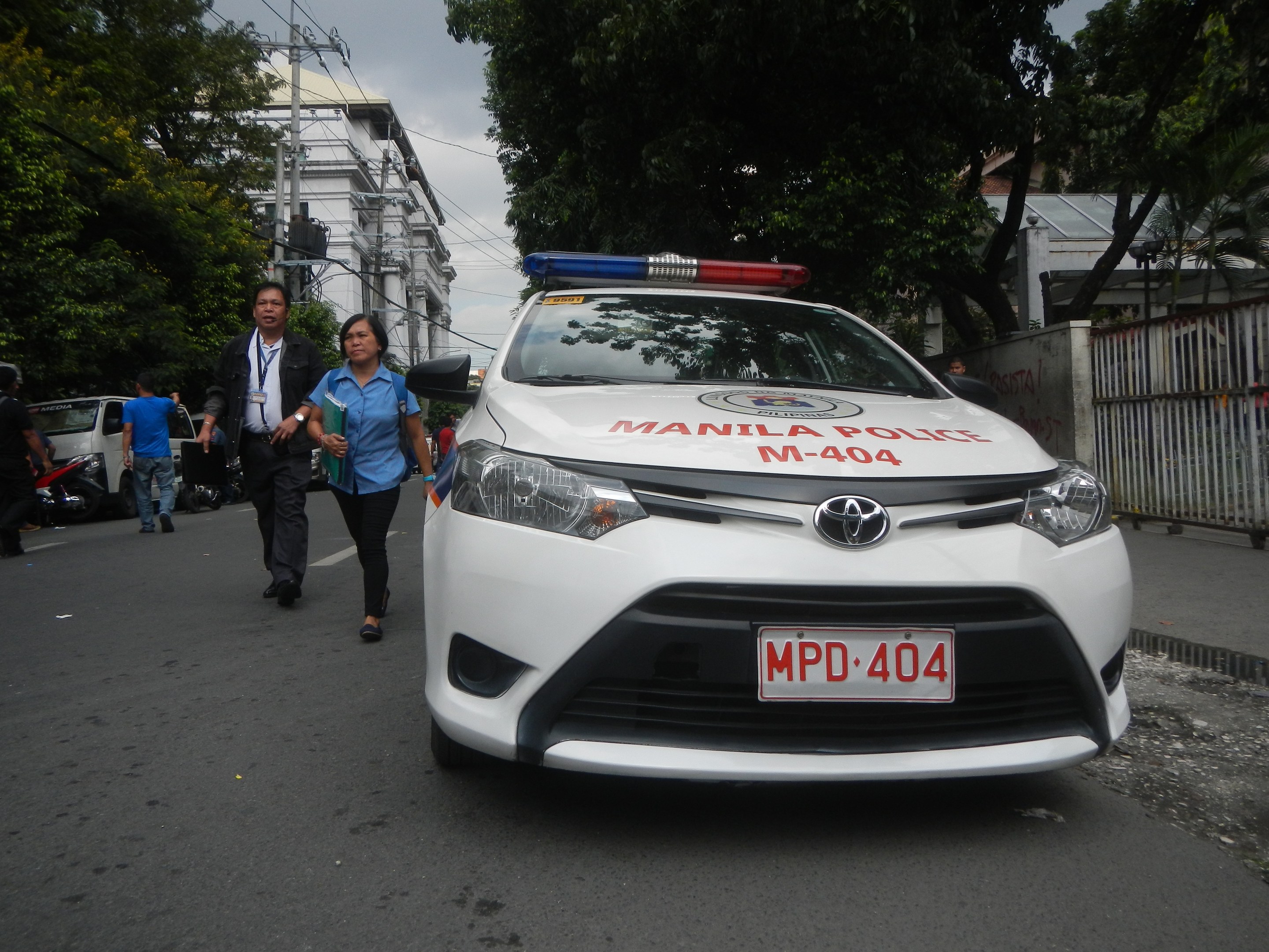 Ein Polizeiwagen steht am Straßenrand in Manila, Philippinen, mit Menschen auf dem Gehweg in der Nähe, einem Geländer und einer Wand auf der rechten Seite und Bäumen, Gebäuden, Pfählen und Drähten im Hintergrund.