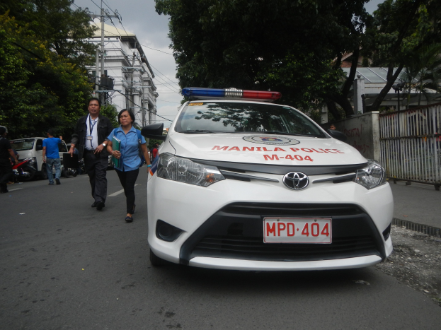 Ein Polizeiwagen steht am Straßenrand in Manila, Philippinen, mit Menschen auf dem Gehweg in der Nähe, einem Geländer und einer Wand auf der rechten Seite und Bäumen, Gebäuden, Pfählen und Drähten im Hintergrund.