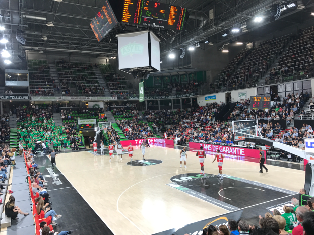 Ein Basketballspiel in einer großen Arena mit Zuschauern in den Rängen, Anzeigetafeln mit "Asvel vs Galatasaray" und einer Decke mit Lampen und einem Bildschirm.