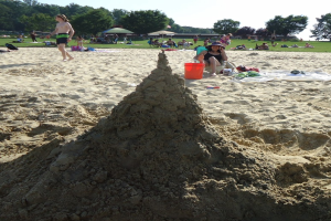 Ein Sandburgenbau am Strand mit spielenden Menschen im Sand, einem Eimer und einer Mütze im Vordergrund und Bäumen und einem klaren blauen Himmel im Hintergrund.
