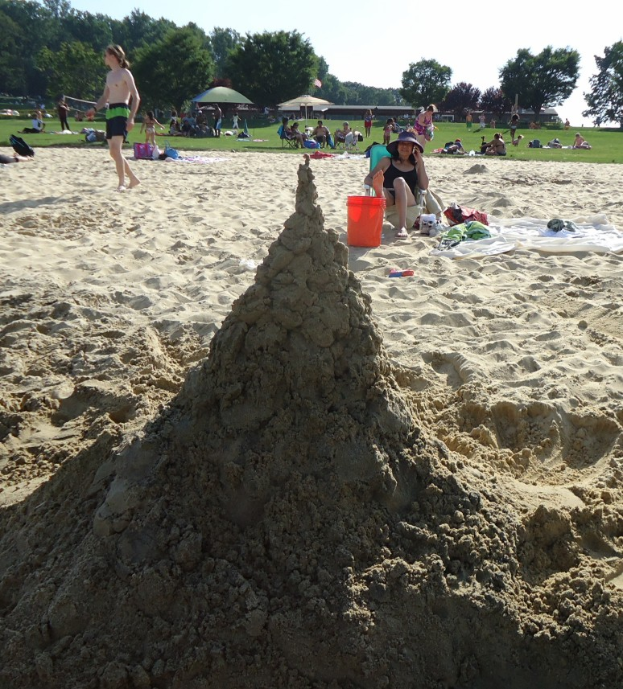 Ein Sandburgenbau am Strand mit spielenden Menschen im Sand, einem Eimer und einer Mütze im Vordergrund und Bäumen und einem klaren blauen Himmel im Hintergrund.