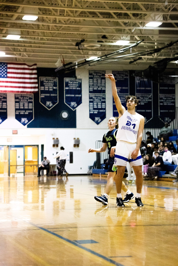 Ein Mann in sportlicher Kleidung wirft einen Basketball in einer Turnhalle, während Zuschauer von Bänken im Hintergrund zusehen.