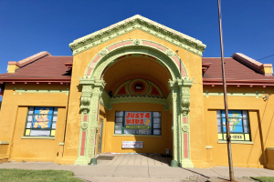 Gelbes Gebäude mit rotem Dach und grünem Dach, beschriftet mit "Just 4 Kids Learning Center", umgeben von Gras und einem Pfahl, mit Bäumen und einem klaren blauen Himmel im Hintergrund.