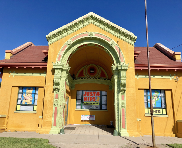 Gelbes Gebäude mit rotem Dach und grünem Dach, beschriftet mit "Just 4 Kids Learning Center", umgeben von Gras und einem Pfahl, mit Bäumen und einem klaren blauen Himmel im Hintergrund.
