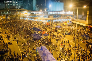 Eine große Menschenmenge steht auf einer nächtlichen Straßenprotest, umgeben von Gebäuden mit Glaswänden, Zelten und einer Brücke im Hintergrund.