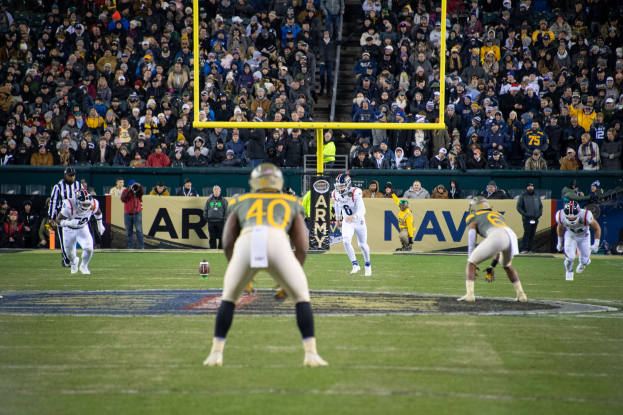 Ein Fußballspiel im Gange mit Spielern in Helmen und Sportbekleidung im Vordergrund und einer Zuschauermenge im Hintergrund, mit sichtbaren Pfählen und Texttafeln.