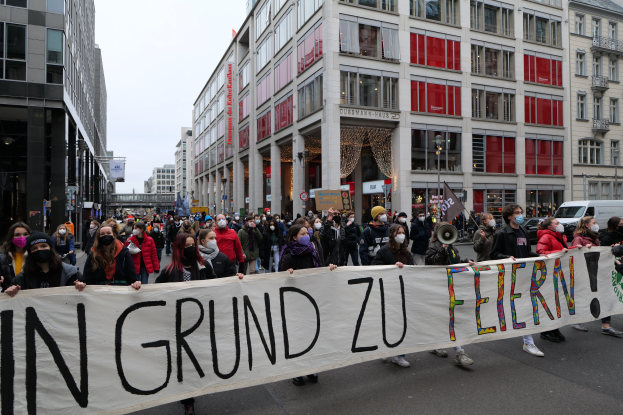 Eine Gruppe von Menschen mit Masken, die ein Transparent mit der Aufschrift "In Grund zu Feiern" tragen, während einer Demonstration in Berlin, Deutschland, mit Gebäuden, Fahrzeugen und Laternenmasten im Hintergrund.