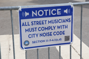 Anschlagtafel mit einem Schild, auf dem "Anschlag: Alle Straßenmusiker müssen die Stadtlärmvorschriften einhalten" steht, an einem Zaun befestigt, mit einer Straße im Hintergrund.