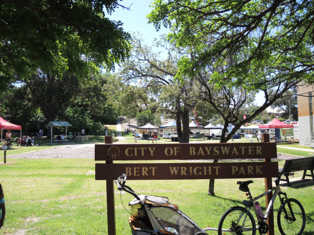 Bayswater Bert Wright Park mit einem Schild, Fahrrad, Kinderwagen, Gras, Bank, Menschen, Zelten, einem Gebäude, einer Straßenlaterne und einem bewölkten Himmel.