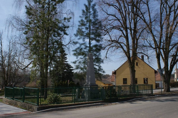 Ein Holocaust-Denkmal steht in der Mitte einer Straße, umgeben von einem Metallzaun, umgeben von Gras, Bäumen, Häusern, einem Straßenmast und einem bewölkten Himmel.