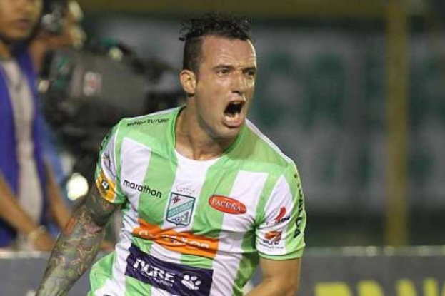 Ein Fußballspieler in grün-weißer Uniform mit Tattoos auf dem Arm feiert nach einem Tor, mit unscharfen Hintergrundfiguren und einer Tafel.
