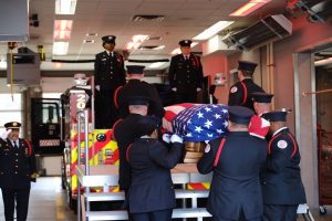 Eine Gruppe uniformierter Männer trägt einen Sarg mit einer amerikanischen Flagge, umgeben von ein paar Männern mit Mützen und einem Feuerwehrauto und Deckenlampen im Hintergrund.