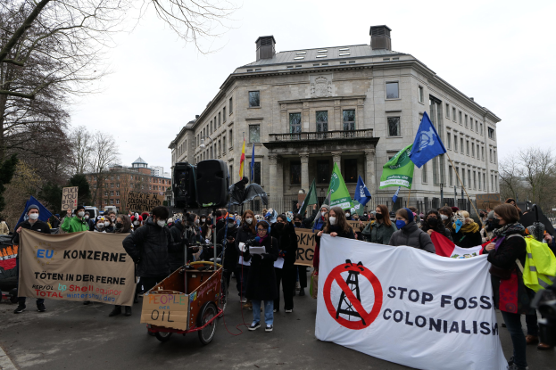 Große Gruppe von Menschen marschiert bei einer Demonstration gegen fossile Brennstoffe, hält Schilder und Fahnen, mit einem Fahrzeug im Vordergrund und Gebäuden, Bäumen und einem klaren blauen Himmel im Hintergrund.